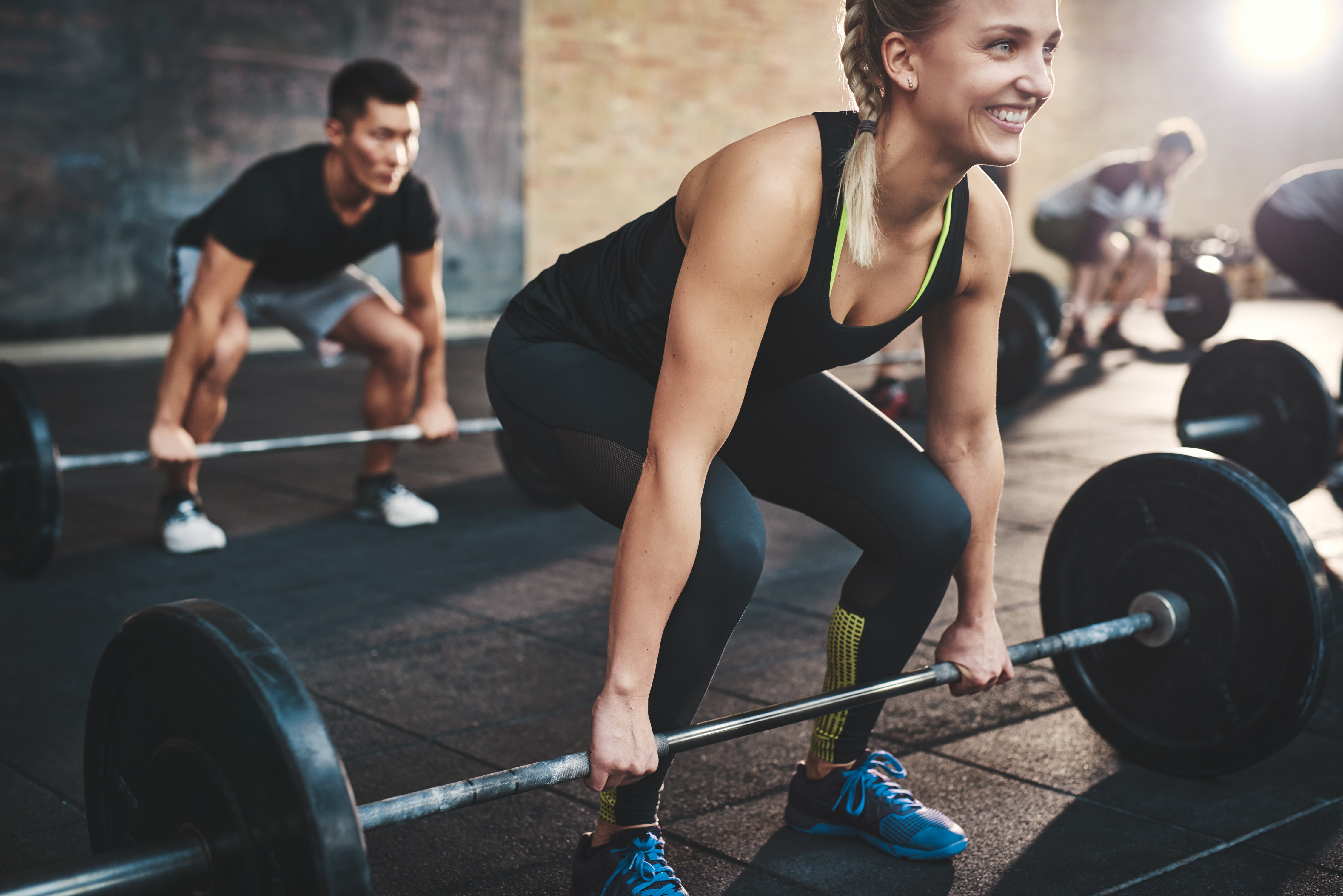 Cheerful muscular young woman with ponytail and black tights performing dead lift barbell exercises with other students